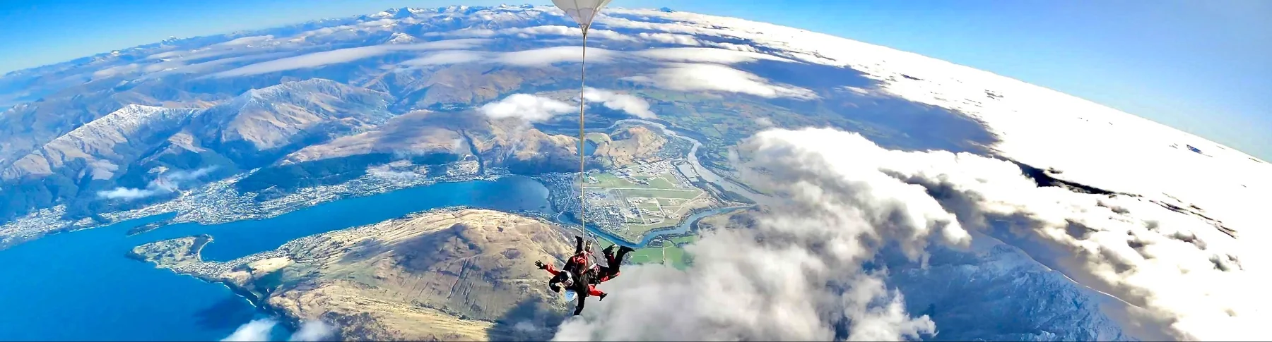 A skydiver above a lake, a town, and snowy mountains.