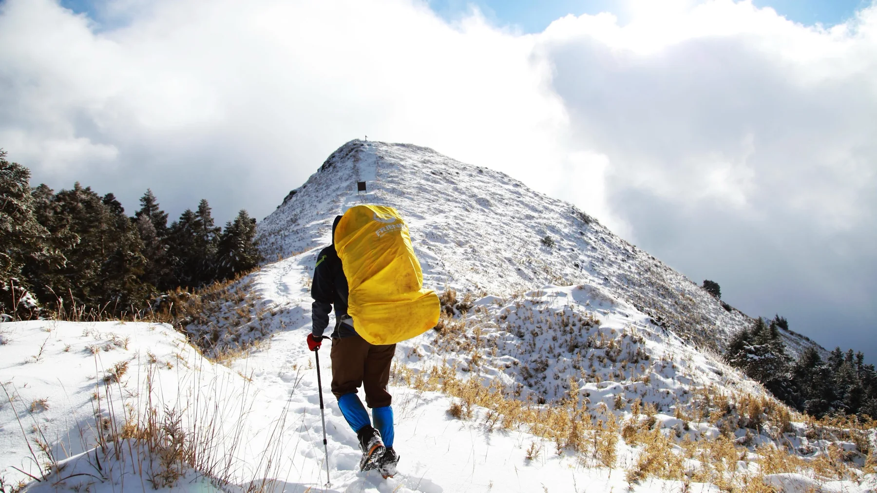 A person with a large yellow pack hiking up a snowy ridge toward a summit.
