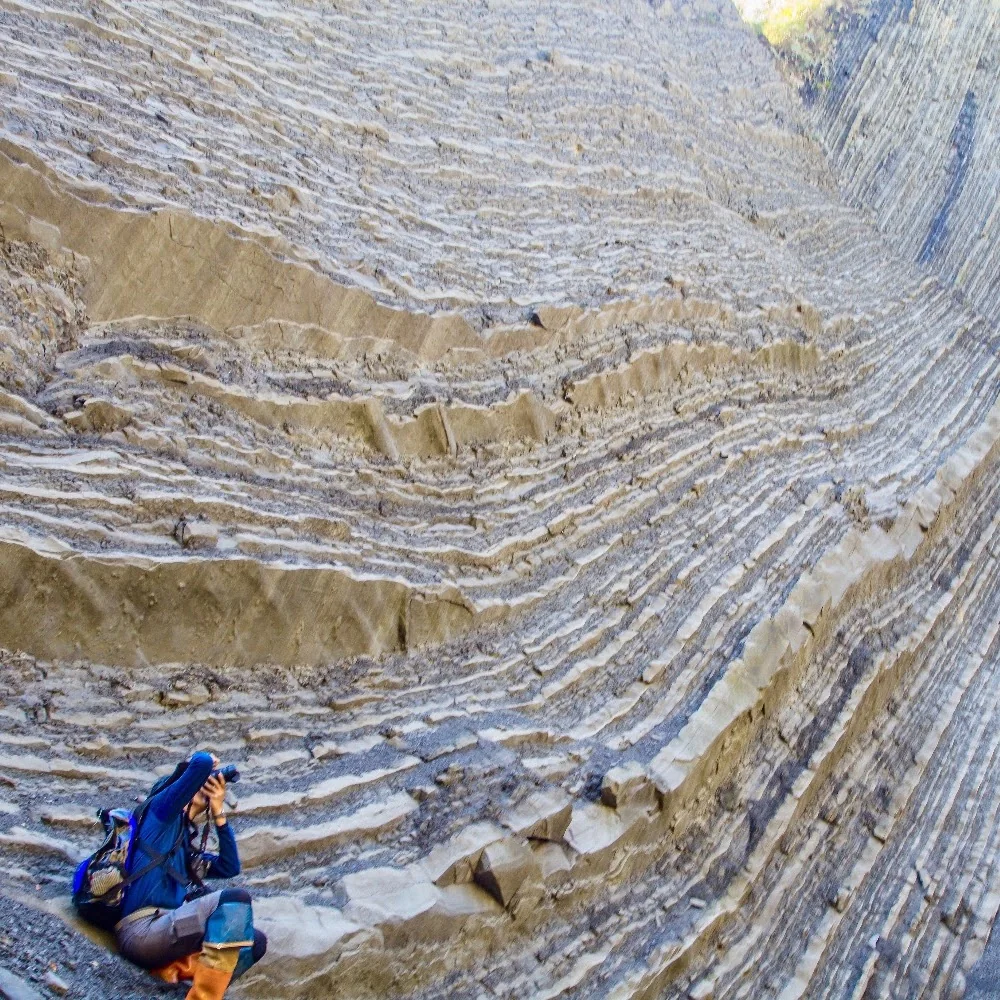 A person photographing a cliff of thin, repeating sedimentary beds.